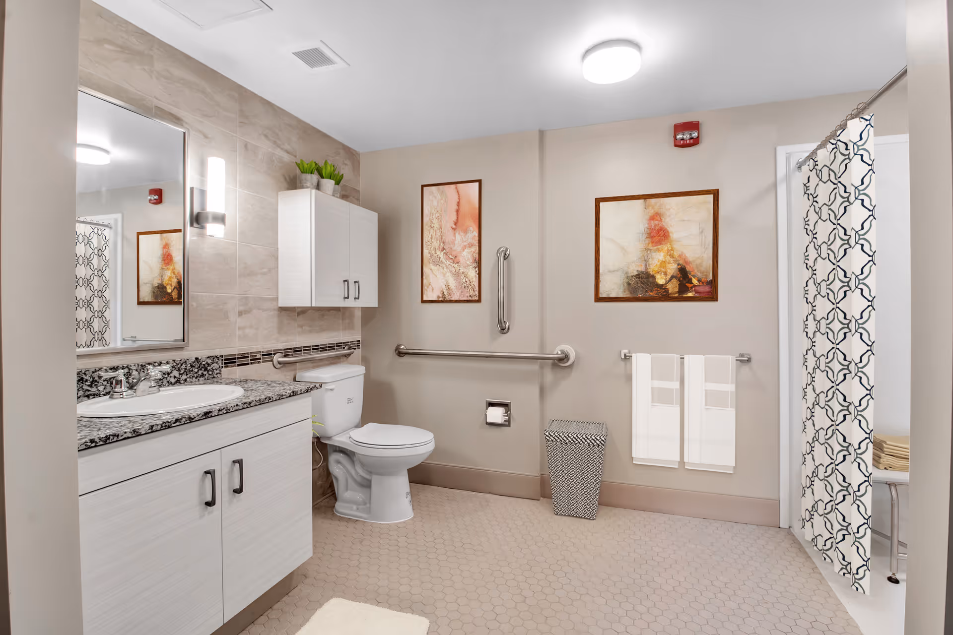 A clean and modern bathroom with a white toilet, a white vanity with granite countertop and sink, a large mirror, and a wall-mounted cabinet. The walls are beige with two framed abstract paintings. There are safety grab bars near the toilet and a patterned shower curtain partially visible on the right. Two white towels hang on a towel rack, and a small wastebasket is on the floor.