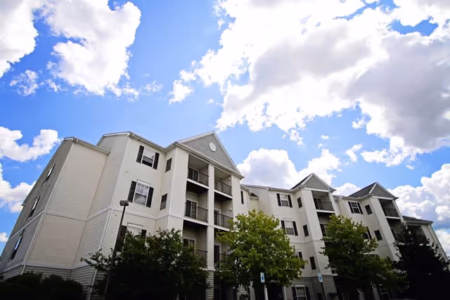 Exterior view of a multi-story residential building with white siding and black window shutters under a partly cloudy blue sky. Trees and greenery are visible in front of the building.