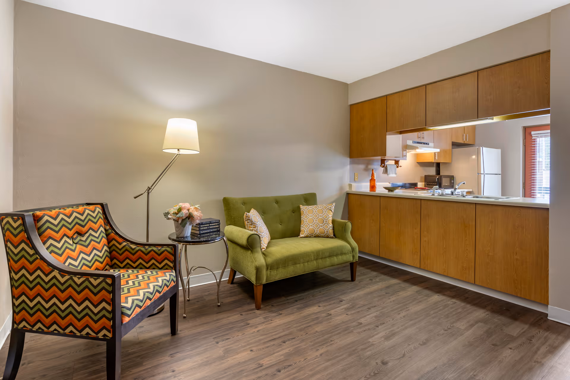 A cozy living area with a green loveseat adorned with two patterned pillows, a colorful zigzag-patterned armchair, a small round side table with a flower arrangement and books, and a floor lamp. The background features a kitchen with wooden cabinets, a white refrigerator, and a countertop with various items.