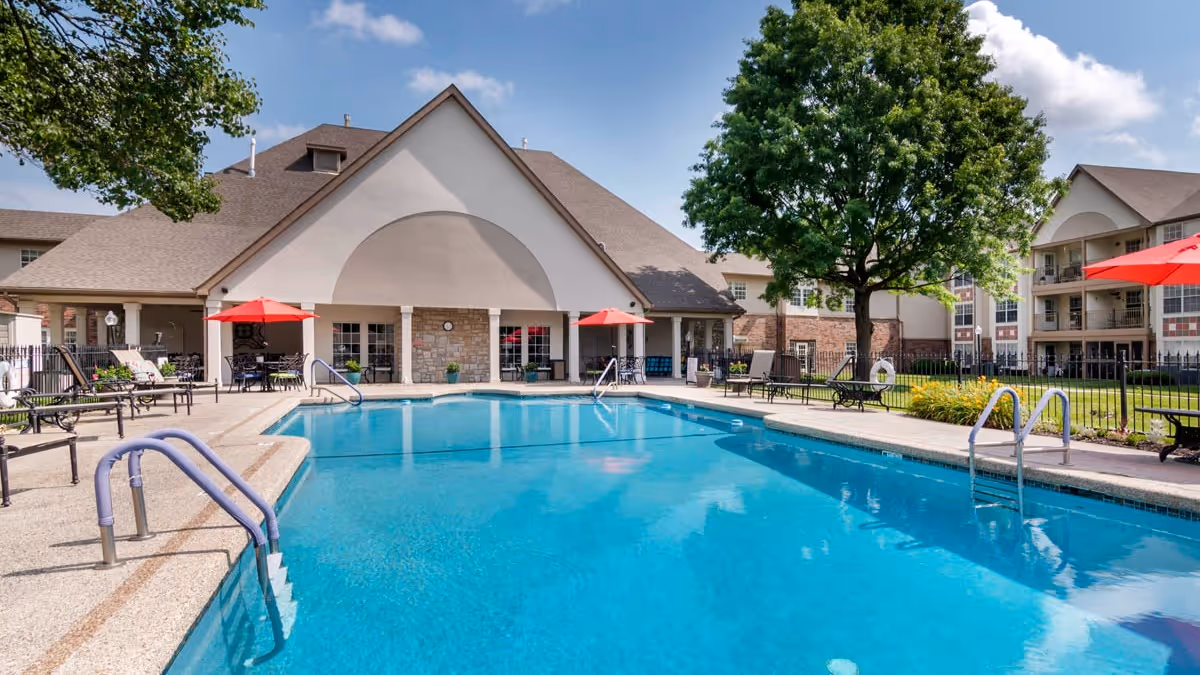 Outdoor swimming pool with lounge chairs, red umbrellas, and a clubhouse building under a blue sky.