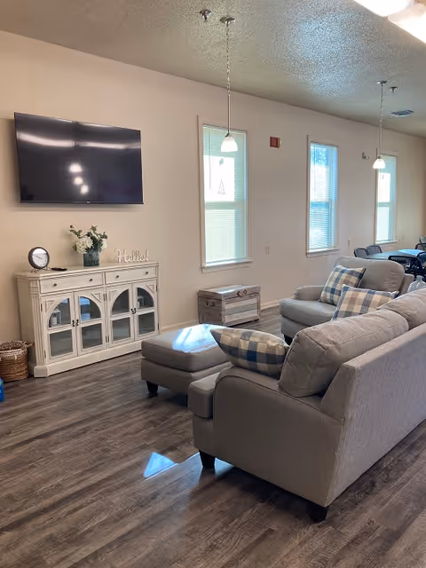 A cozy living room area with a gray sofa, matching armchair, and ottoman, all adorned with blue and white checkered pillows. A white cabinet with glass doors holds decorative items including a clock, a vase with flowers, and a 'Hello!' sign. A flat-screen TV is mounted on the wall above the cabinet. The room has wood-style flooring, three windows with blinds, and pendant lights hanging from the ceiling. In the background, a dining table with chairs is partially visible.