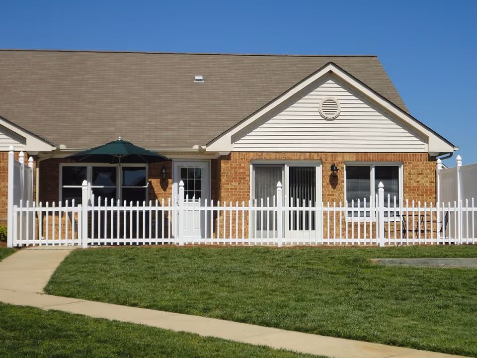 Single-story brick building with a beige roof and white trim, featuring a white picket fence enclosing a small patio area with a green umbrella and outdoor chairs. A concrete walkway curves in front of the building, and the sky is clear and blue.