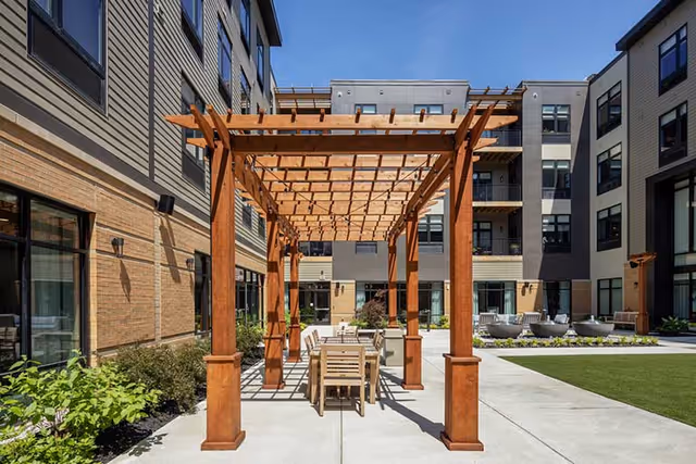 Outdoor courtyard area at The Summit of Blue Ash featuring a wooden pergola with a dining table and chairs underneath. The courtyard is surrounded by a multi-story building with large windows, landscaped greenery, and a clear blue sky overhead.