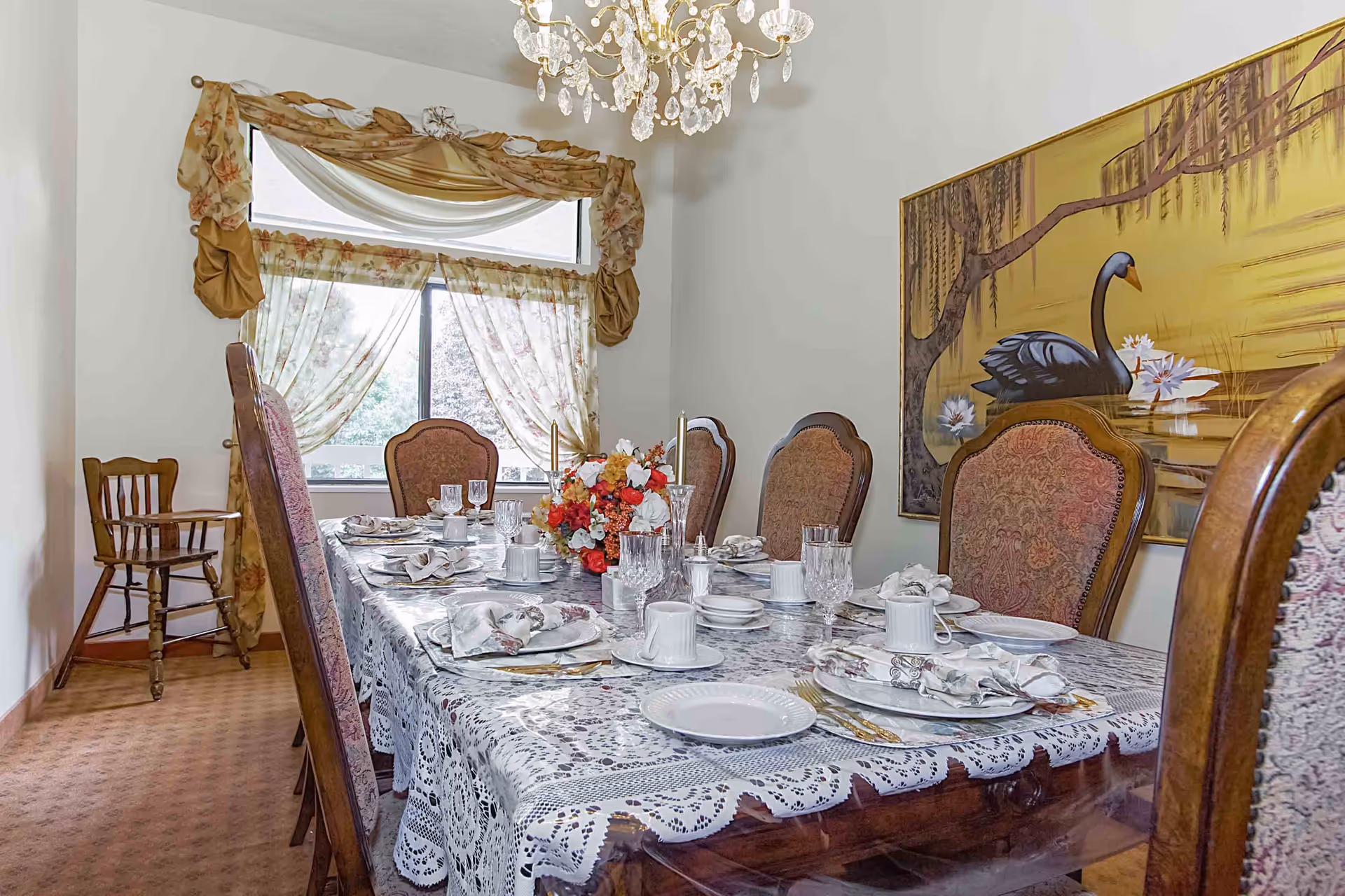 A formal dining room with a long wooden table covered by a lace tablecloth, set with plates, cups, glasses, and napkins. The room features upholstered dining chairs, a floral centerpiece, a chandelier hanging from the ceiling, a window with floral curtains, and a large painting of a black swan on the wall.