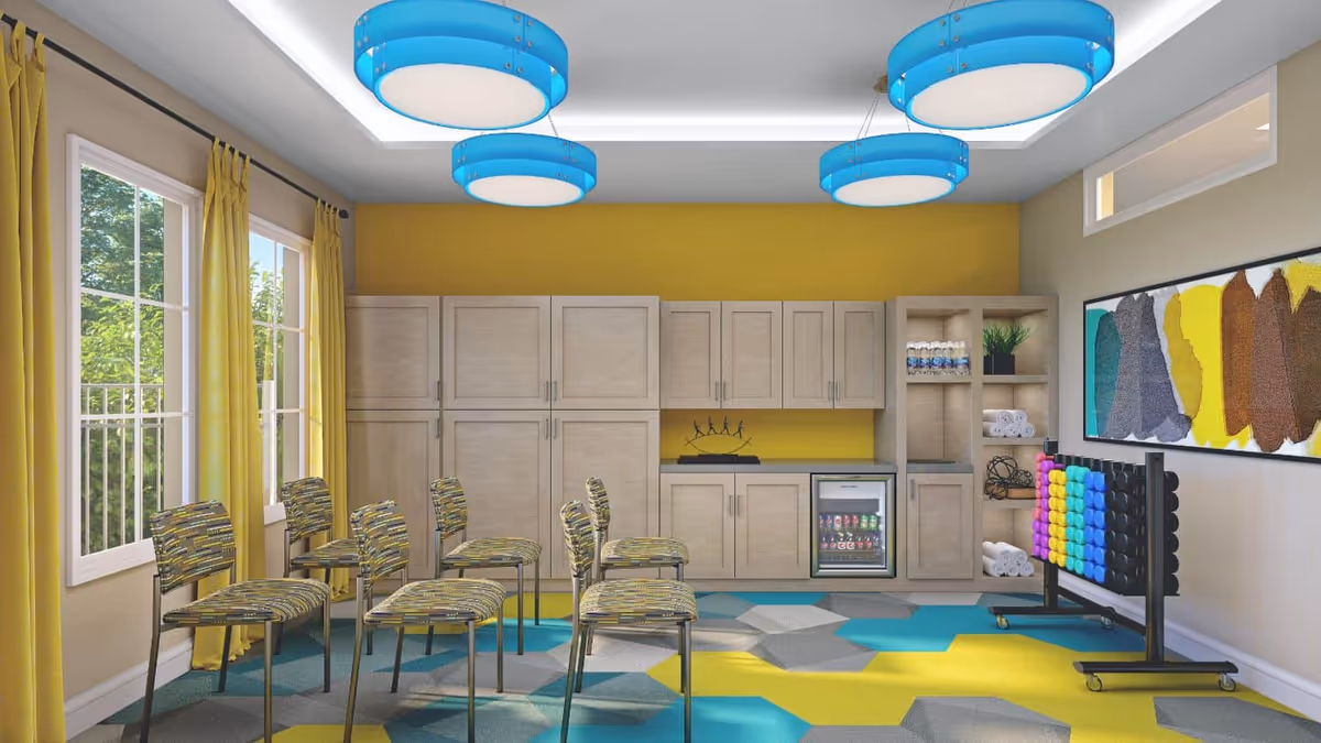 A bright, modern room with yellow walls and large windows with yellow curtains. The room features a set of six patterned chairs arranged in two rows, a wall of light wood cabinets with a small refrigerator stocked with drinks, and open shelving holding towels, water bottles, and a plant. The floor has a colorful geometric pattern in blue, yellow, and gray. Blue circular ceiling lights hang from the ceiling, and a large abstract painting with yellow, brown, and teal colors is on the wall. A large Connect Four game with colorful discs is also present.
