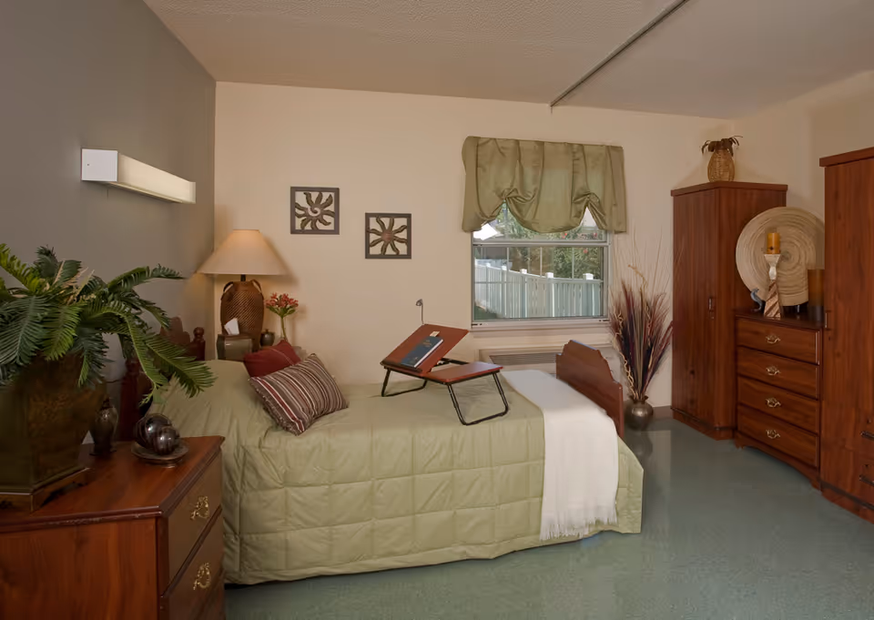 A cozy bedroom in a senior living facility with a single bed covered in a green quilt and a white throw blanket. The bed has a striped pillow and a wooden adjustable tray on top. Next to the bed is a wooden nightstand with a large lamp, a small vase with flowers, and decorative items. There is a window with a green valance, and wooden wardrobes and dressers with decorative items on top are placed against the walls. A potted plant is on another wooden dresser in the foreground.