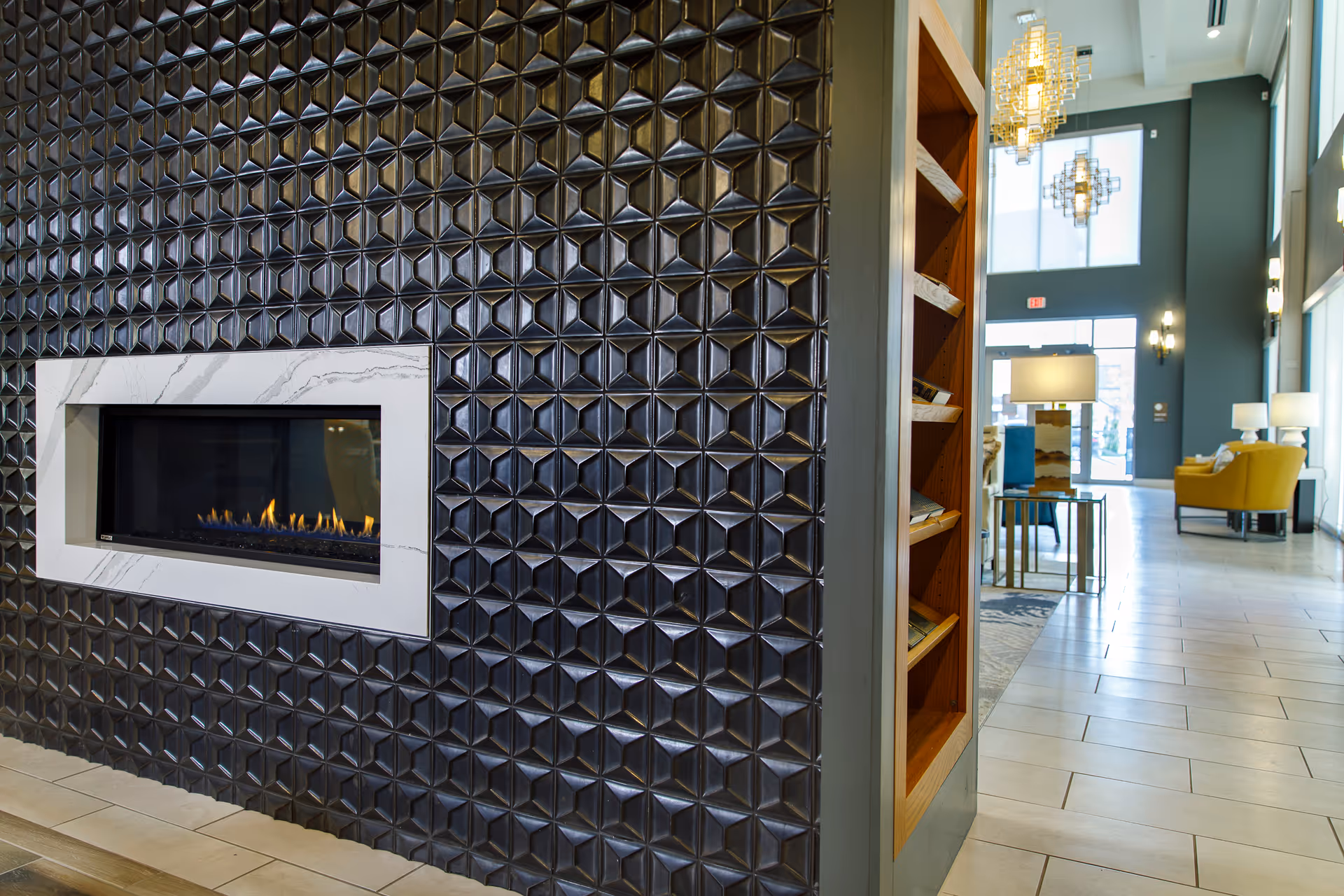 A modern interior space featuring a decorative black tiled wall with a built-in rectangular gas fireplace. To the right, there is a tall bookshelf with magazines or brochures, and further in the background, a seating area with yellow chairs, tables, and large windows letting in natural light.