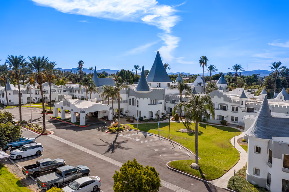 Aerial view of Camelot - A Provincial Senior Living Community featuring white castle-like buildings with blue conical roofs, surrounded by palm trees, green lawns, and a parking lot with several cars under a clear blue sky.
