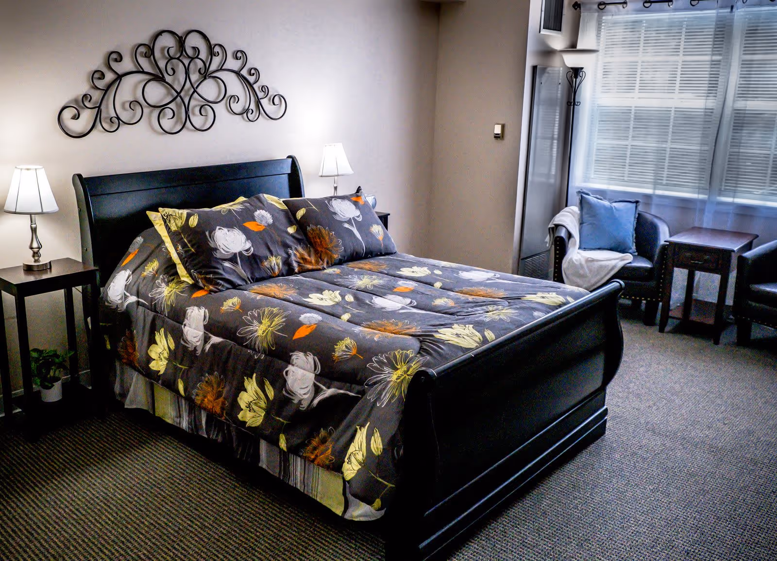A cozy bedroom in Cherry Hill Senior Living featuring a black wooden bed with floral bedding, two bedside tables with lamps, a decorative metal wall art above the bed, a window with blinds and sheer curtains, and two armchairs with a small table between them.