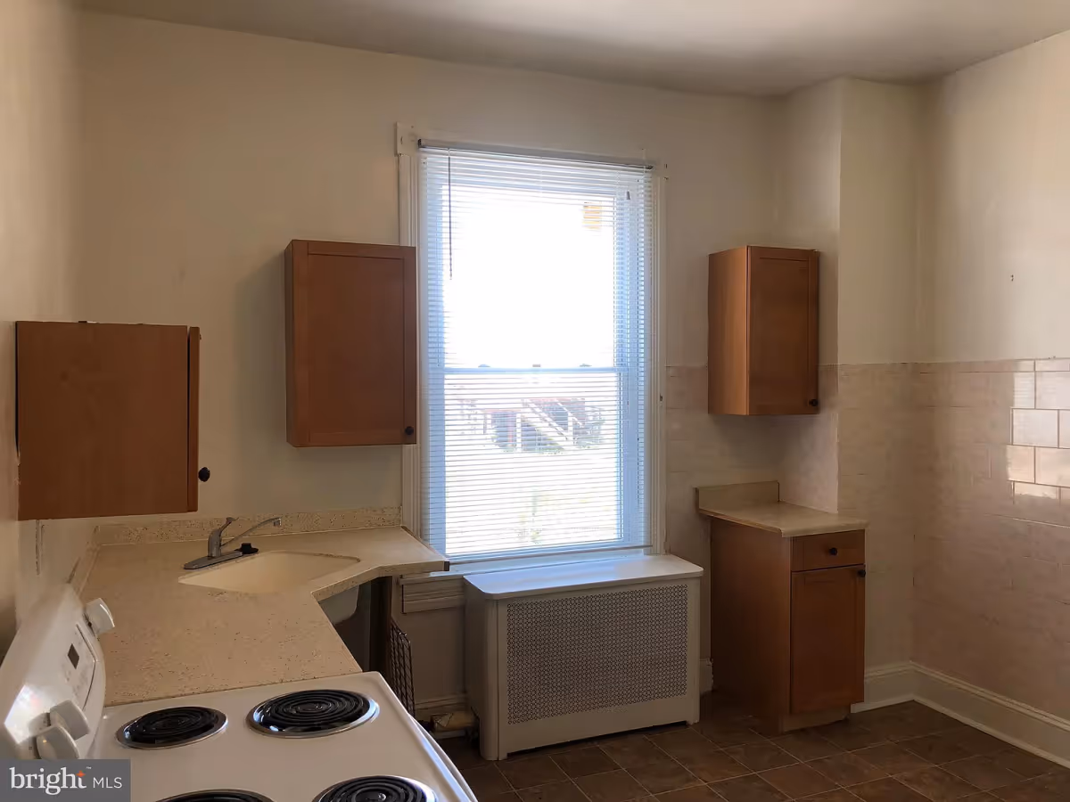 A small kitchen with a white electric stove, beige countertops, wooden cabinets mounted on the walls, a window with white blinds, and beige tiled walls and floor.