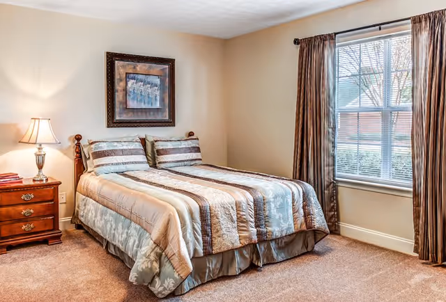 A cozy bedroom with a neatly made bed featuring a striped comforter and matching pillows. There is a wooden nightstand with a lamp and books on the left side of the bed. A framed artwork hangs on the wall above the bed. A large window with brown curtains allows natural light to brighten the room.