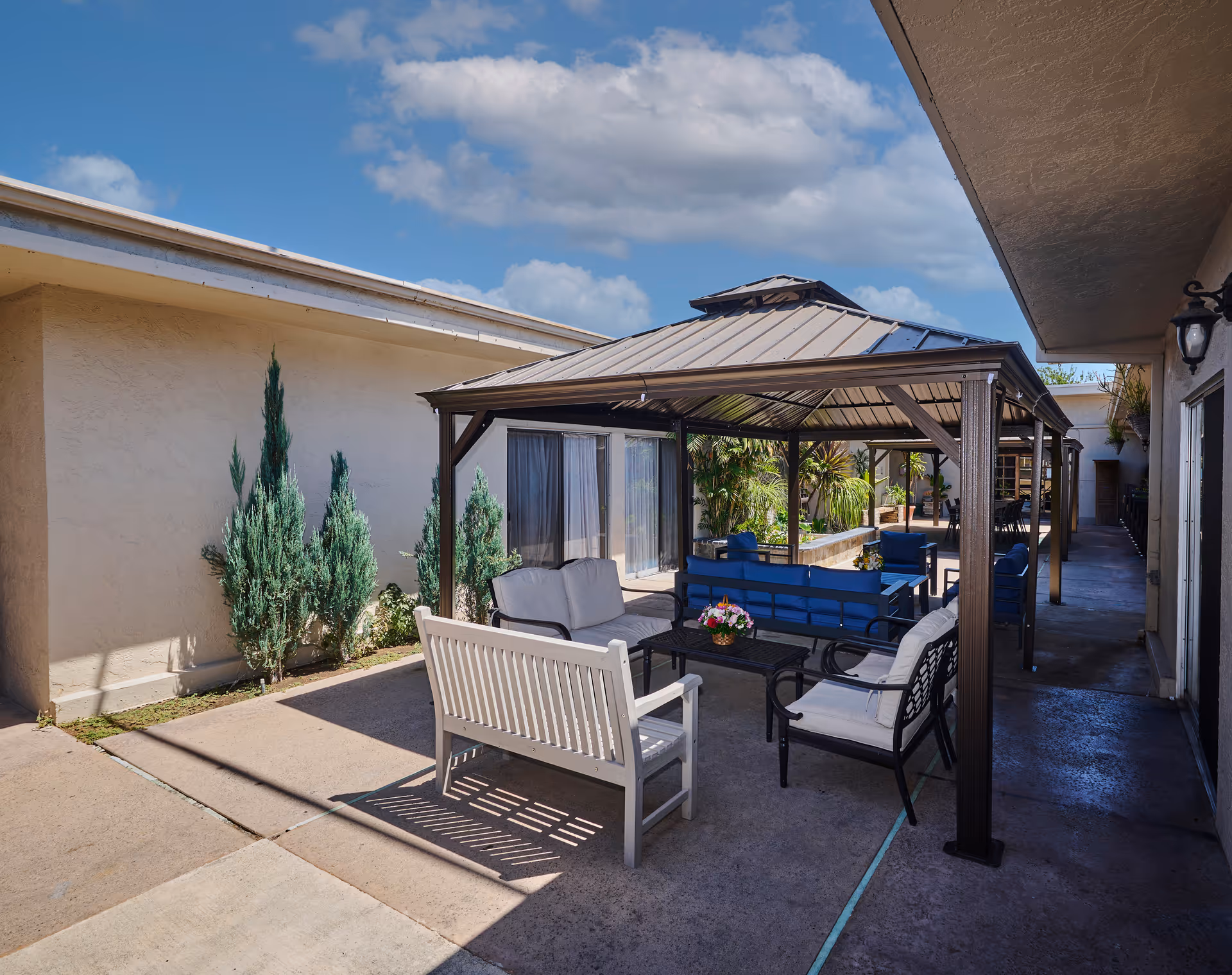 Outdoor patio area at Huntington Park Nursing Center featuring a metal gazebo with seating underneath, including white and blue cushioned chairs and benches, surrounded by plants and adjacent to the building walls under a partly cloudy sky.