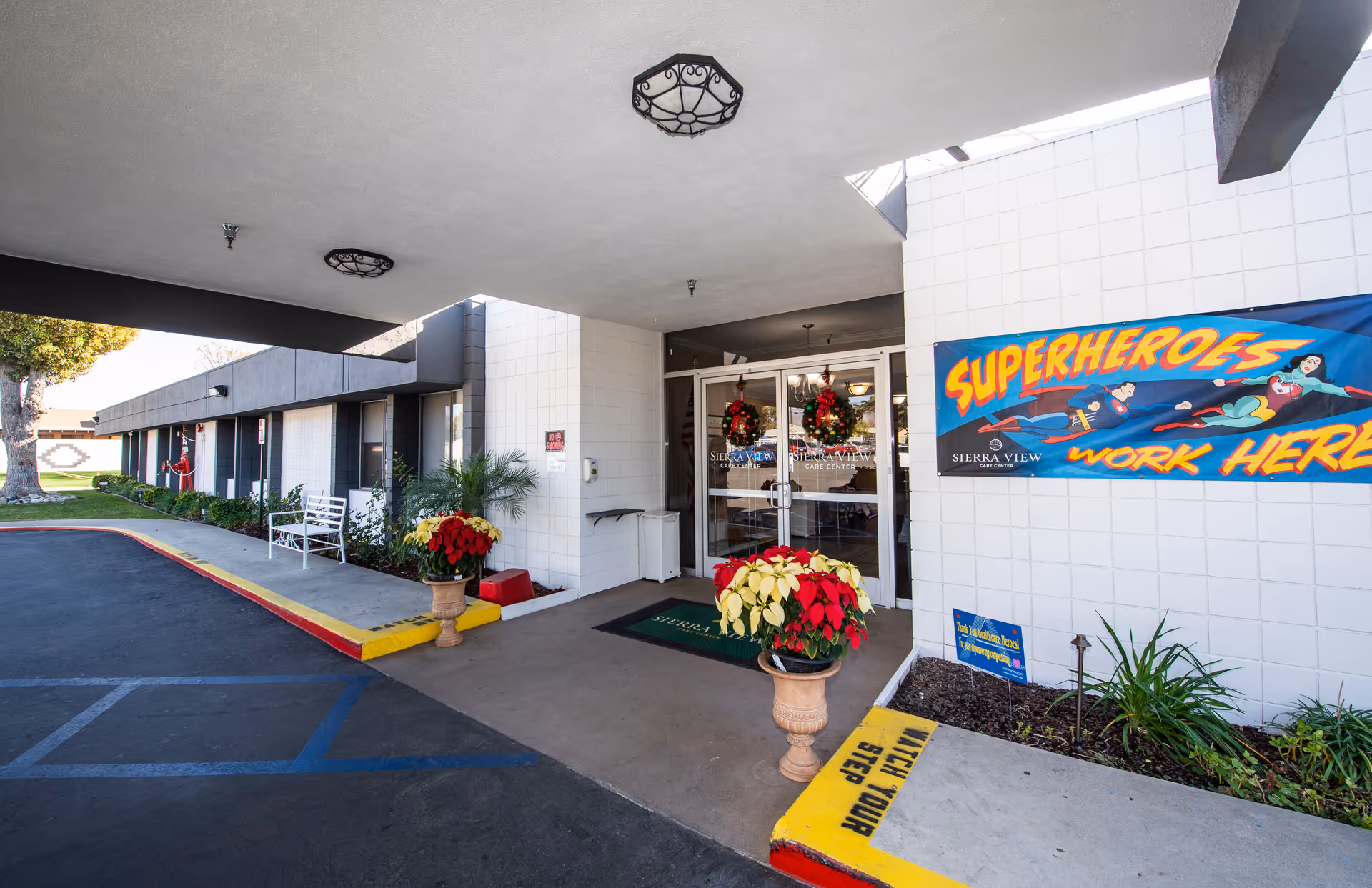 Covered main entrance of Sierra View Care Center with glass double doors, holiday wreaths and poinsettia planters, a 'Superheroes work here' banner, and a drive-up drop-off area.