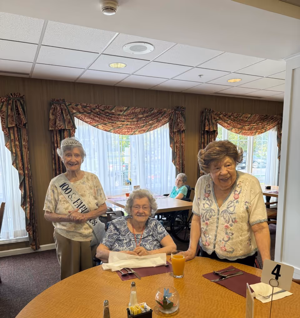 Three smiling women gathered around a set dining table in a cozy, curtain‑draped dining room.