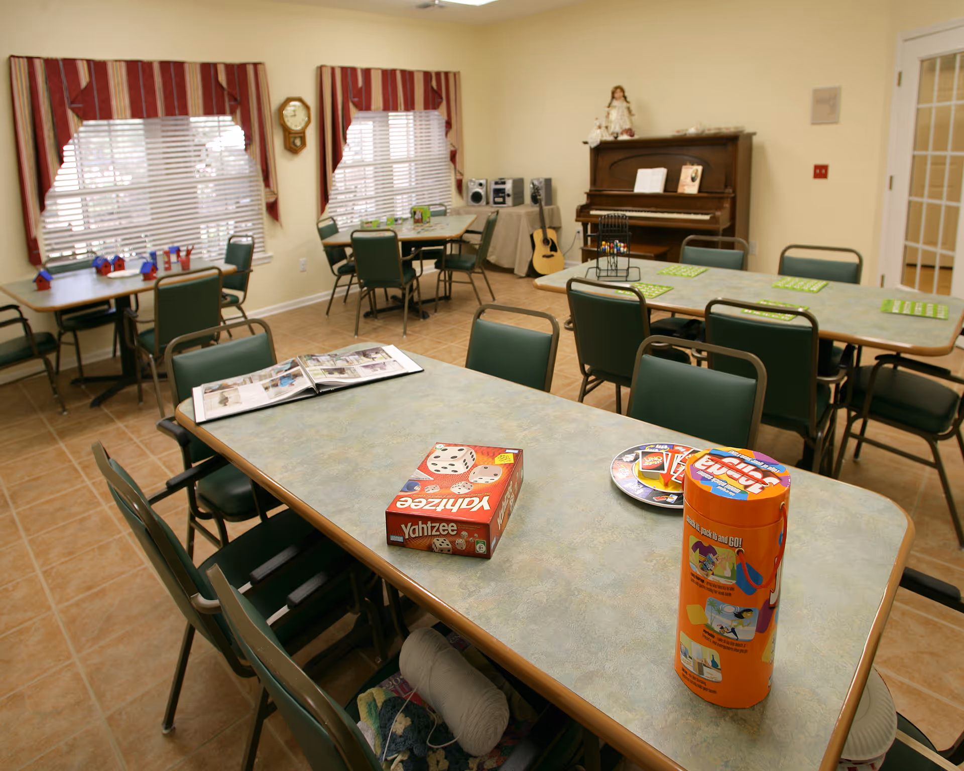 A senior living facility common room with several tables and green chairs arranged around them. On the tables are board games including Yahtzee and other game pieces. In the background, there is a piano with a doll on top, a guitar leaning against the wall, and a small stereo system. The room has tiled floors, two windows with striped red and beige valances, and a wall clock between the windows.