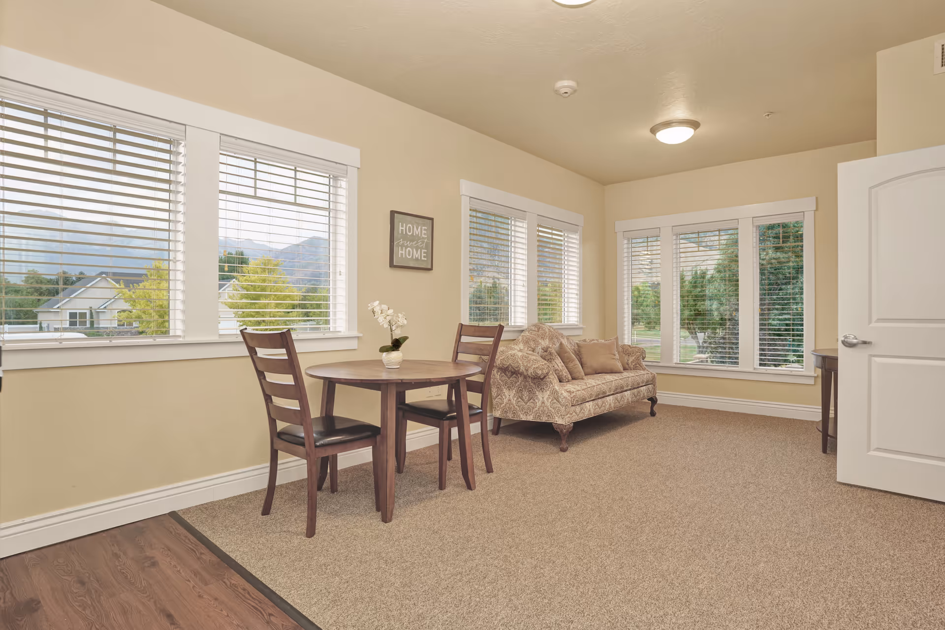 A bright living room area with beige walls and carpeted floor. There is a small round wooden table with two chairs, a patterned sofa with cushions, and multiple large windows with white blinds letting in natural light. A small decorative sign on the wall reads 'HOME sweet HOME'.