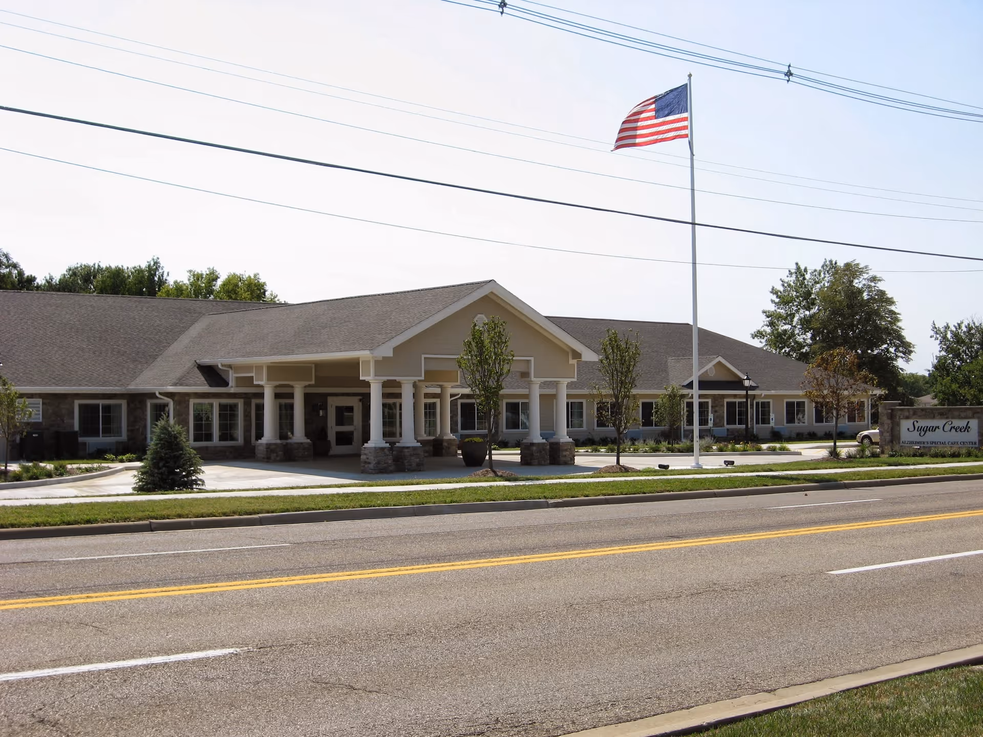 Exterior view of Sugar Creek Alzheimer's Special Care Center, a single-story building with a covered entrance supported by columns, an American flag on a tall flagpole, and a sign displaying the facility's name. The building is surrounded by small trees and landscaping, with a road in the foreground.