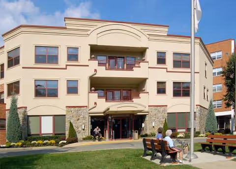 Front view of a beige senior living building with entrance, balconies, and benches.
