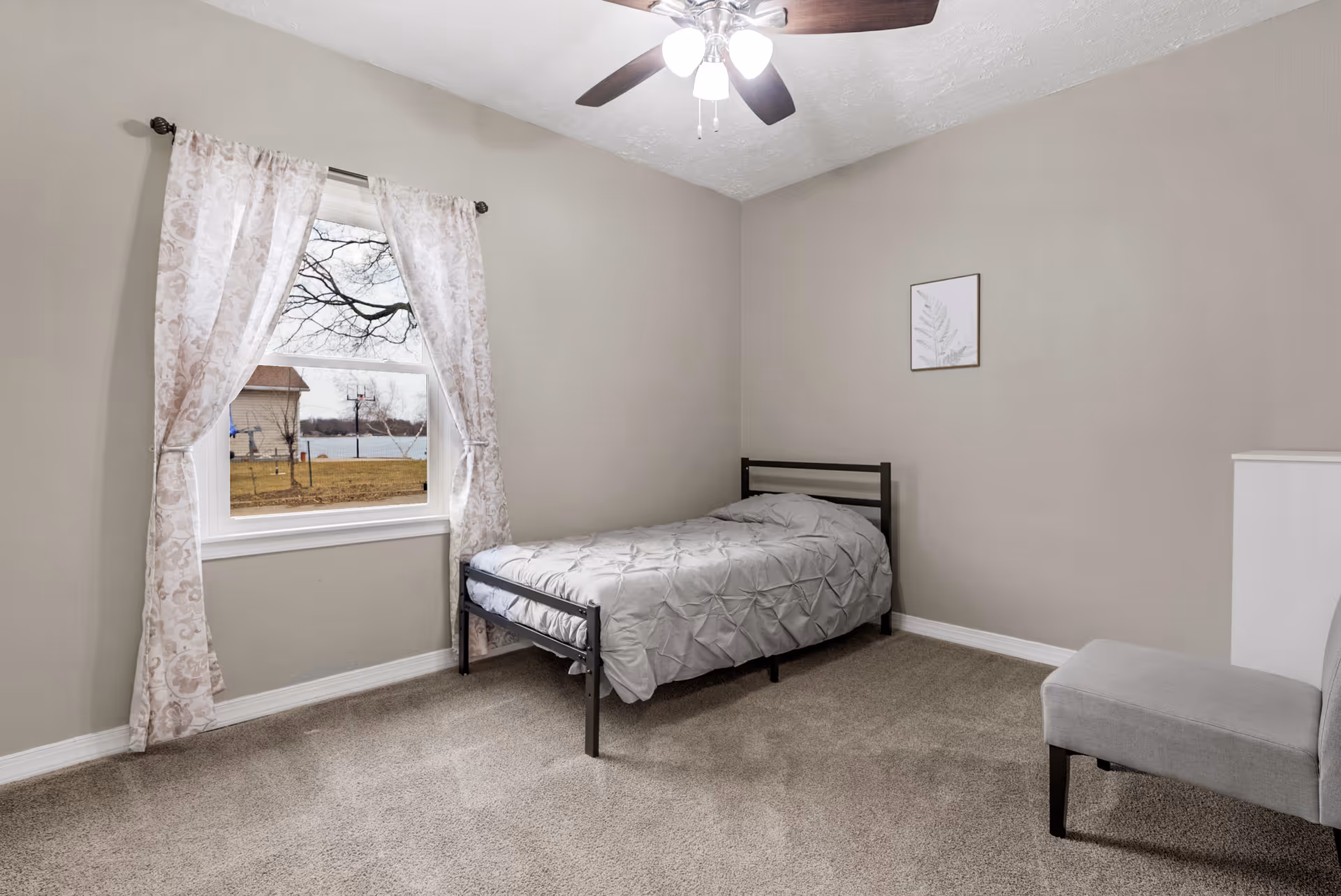 A simple bedroom with a single bed covered in a gray comforter, a gray upholstered chair, beige walls, a window with floral curtains showing an outdoor view, and a ceiling fan with lights.