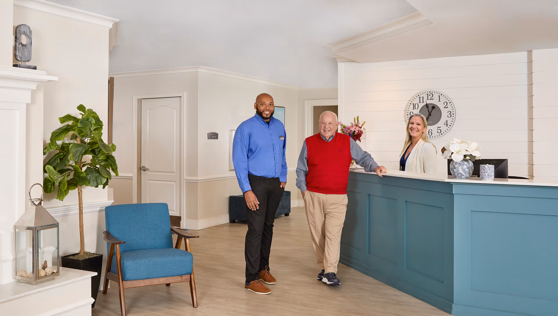 A senior man and a staff member stand smiling in a bright reception area of a senior living facility. A woman is behind the blue reception desk, also smiling. The room features light-colored walls, a large clock on the wall, a blue armchair, a potted plant, and decorative items on the desk and mantel.