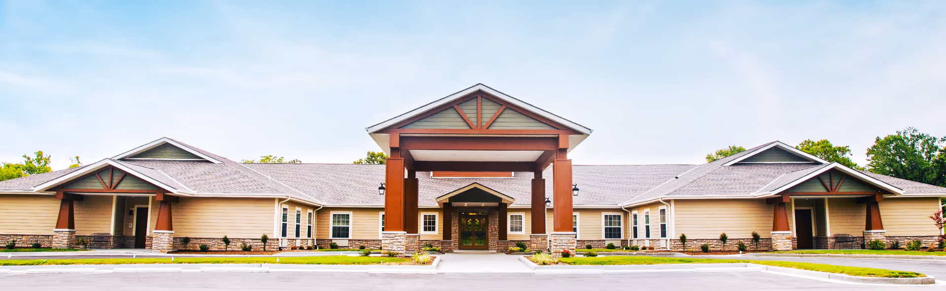 Front exterior view of Oak Pointe of Rolla, a single-story building with a large covered entrance supported by wooden beams and stone pillars, surrounded by a well-maintained lawn and parking area under a clear sky.