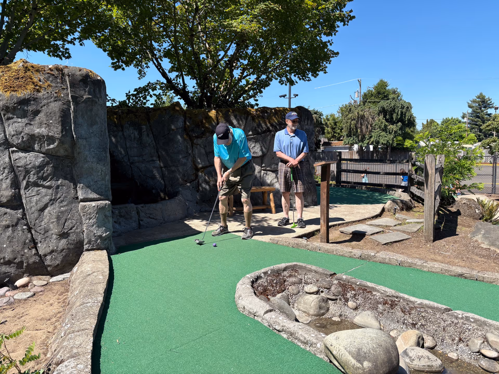 Two elderly men playing mini golf on a green putting surface surrounded by large artificial rock formations and landscaping under a clear blue sky.