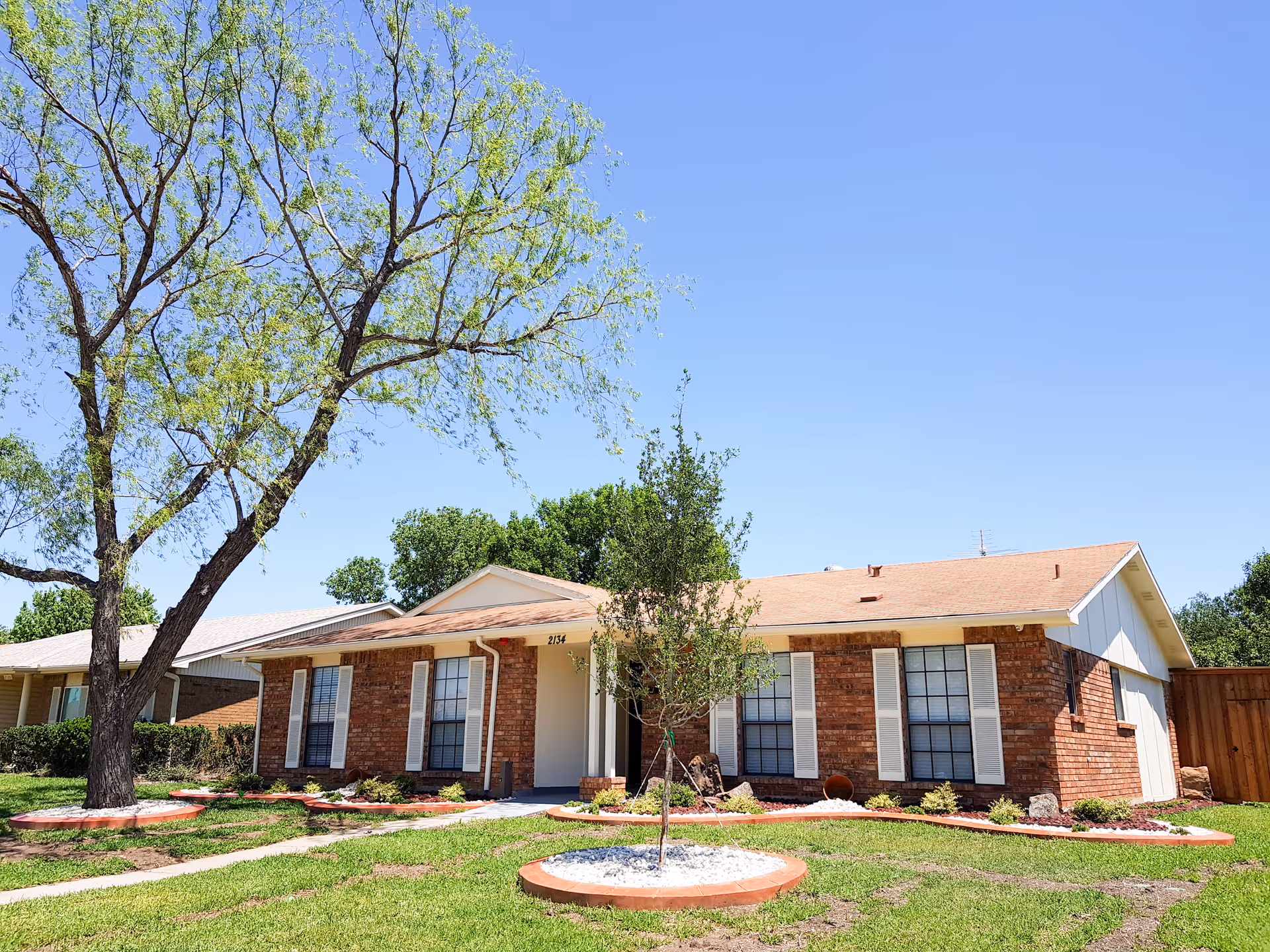 Single-story brick house with white window shutters and a light brown roof, surrounded by a green lawn with a few small trees and a larger tree on the left side under a clear blue sky.