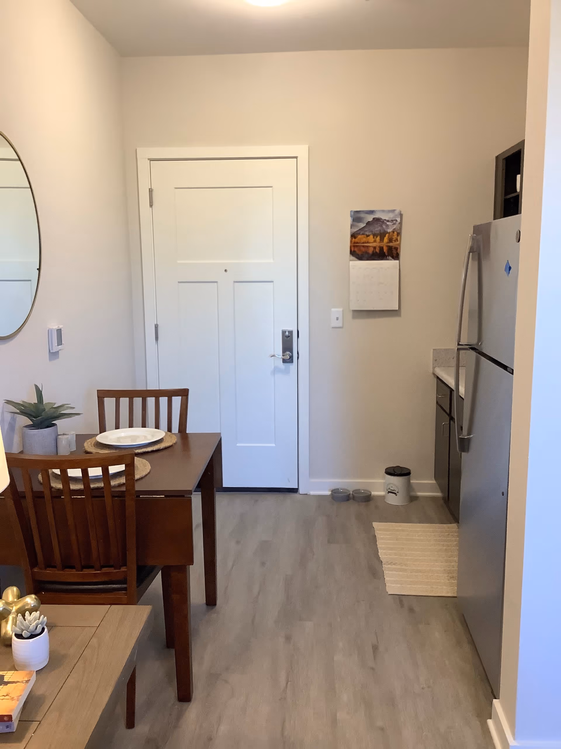 Small apartment entry and kitchenette with a wooden dining table and chairs, a stainless steel refrigerator, and a closed white door.