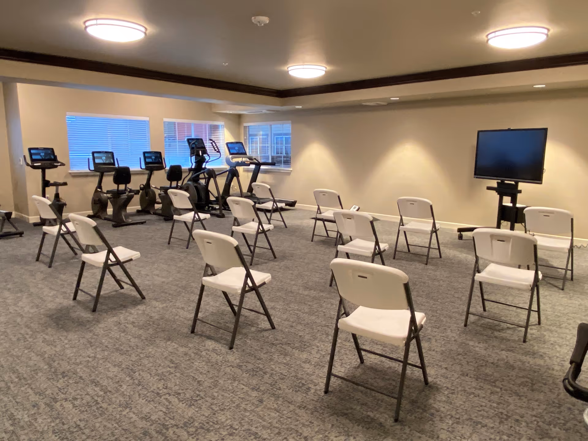 A fitness or activity room with several white folding chairs arranged in rows facing a large flat-screen TV on a stand. In the background, there are exercise machines including stationary bikes and elliptical trainers near windows with blinds. The room has carpeted flooring and ceiling lights.