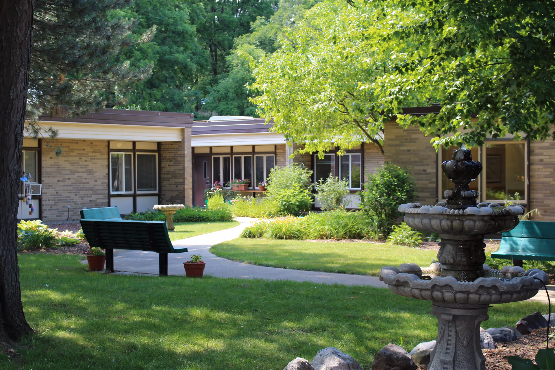 Outdoor garden area at MediLodge of Grand Rapids featuring green grass, trees, shrubs, a stone fountain, two green benches, and a paved walkway surrounded by a single-story brick building.