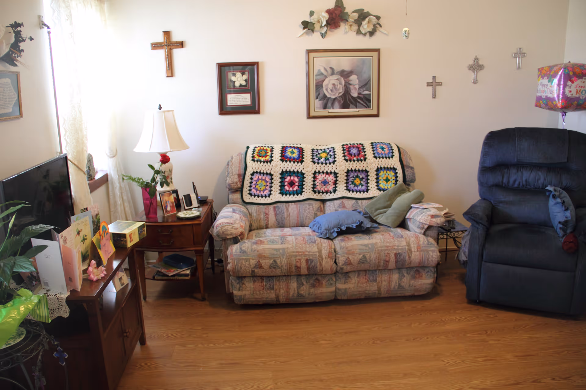A cozy living room with a patterned loveseat covered by a colorful crocheted blanket, a dark recliner chair, and a wooden side table with a lamp and framed photos. The walls are decorated with several crosses and framed floral artwork. A television and various greeting cards are on a wooden TV stand near a window with sheer curtains.