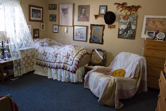 A cozy bedroom with a single bed covered in a floral quilt, a cushioned armchair draped with a blanket, and a wooden dresser. The walls are decorated with various framed pictures and artwork, including a portrait of Jesus and images of horses. A window with lace curtains lets in natural light.