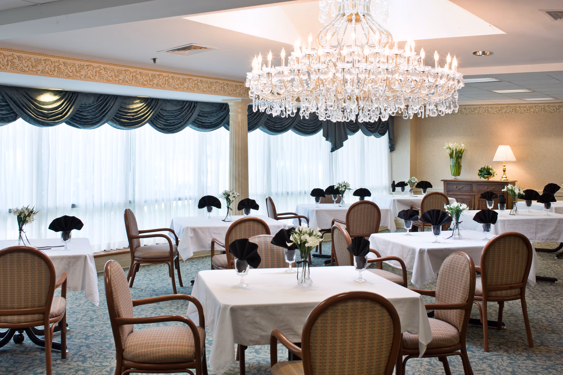 A bright dining room with multiple tables covered in white tablecloths, each set with black folded napkins and small flower arrangements. The room features large windows with sheer white curtains and dark green valances, a large ornate chandelier hanging from the ceiling, and a sideboard with a lamp and additional flower arrangements.