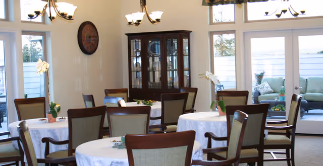A bright dining room with round tables covered in white tablecloths, surrounded by wooden chairs with cushioned seats. A dark wooden china cabinet is against the wall, and a large clock hangs above it. There are two chandeliers hanging from the ceiling, and large windows and glass doors let in natural light, showing an outdoor seating area with cushioned chairs and a small table.