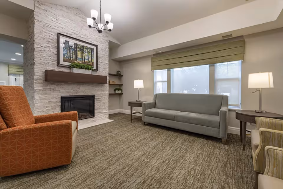 A cozy living room with a gray sofa, an orange armchair, two round wooden side tables each with a lamp, and a fireplace with a stone surround. Above the fireplace is a framed picture of a forest scene and a small plant on the mantel. The room has large windows with green Roman shades and beige walls.