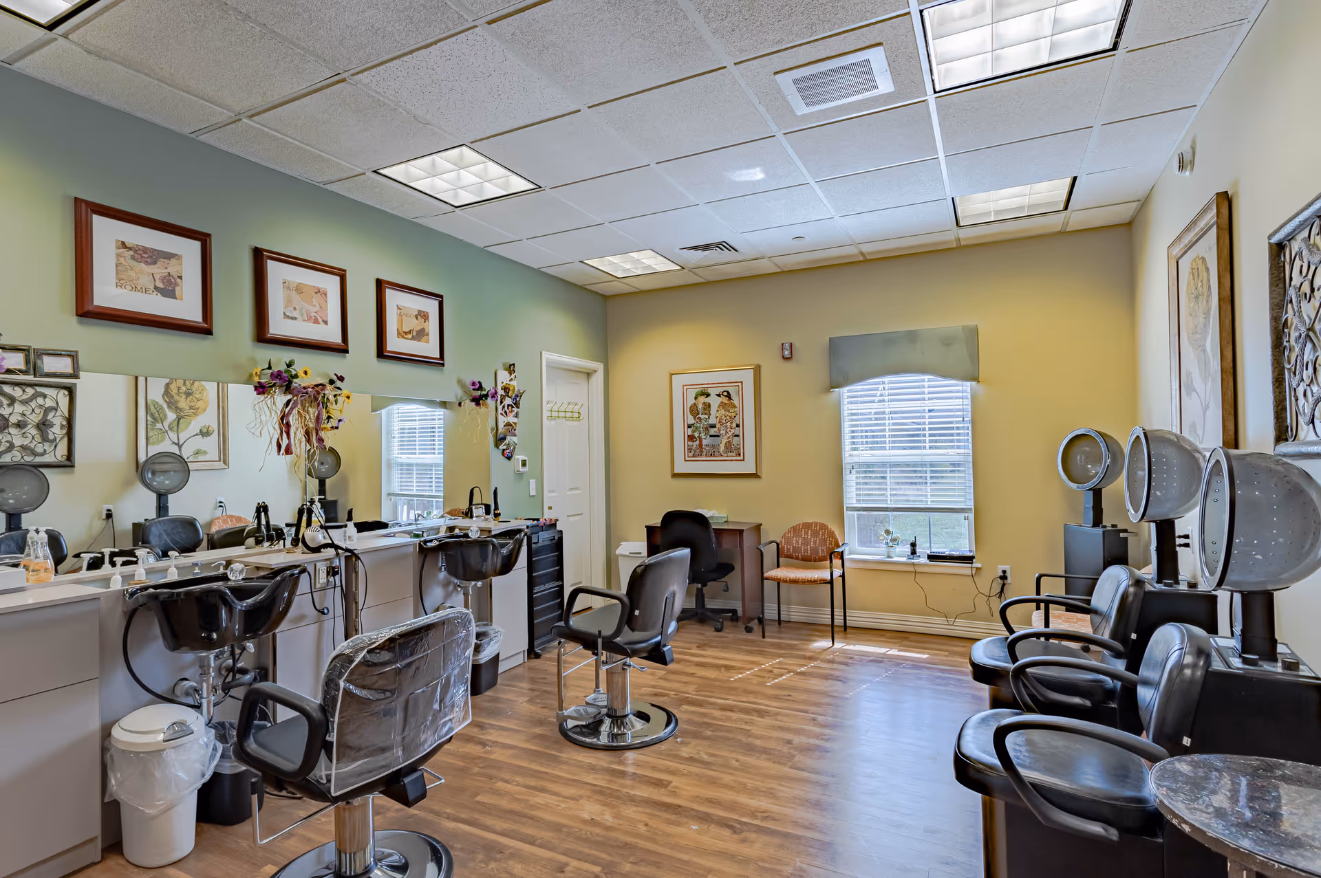 A bright and clean hair salon area inside Bear Creek Assisted & Senior Living facility. The room features multiple salon chairs in front of mirrors with hair dryers mounted on the wall. The walls are decorated with framed artwork and floral arrangements. There is a window with blinds letting in natural light, and the floor is wooden.