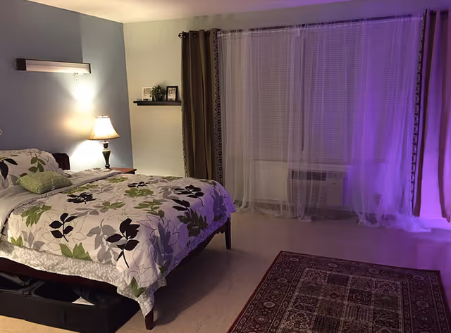 A cozy bedroom with a bed covered in a floral patterned bedspread in shades of green, black, and gray. There is a nightstand with a lamp beside the bed, a small wall shelf with framed pictures and a plant, and a large window with sheer white curtains and darker drapes. A patterned area rug is on the floor near the window, and soft purple lighting illuminates the right side of the room.