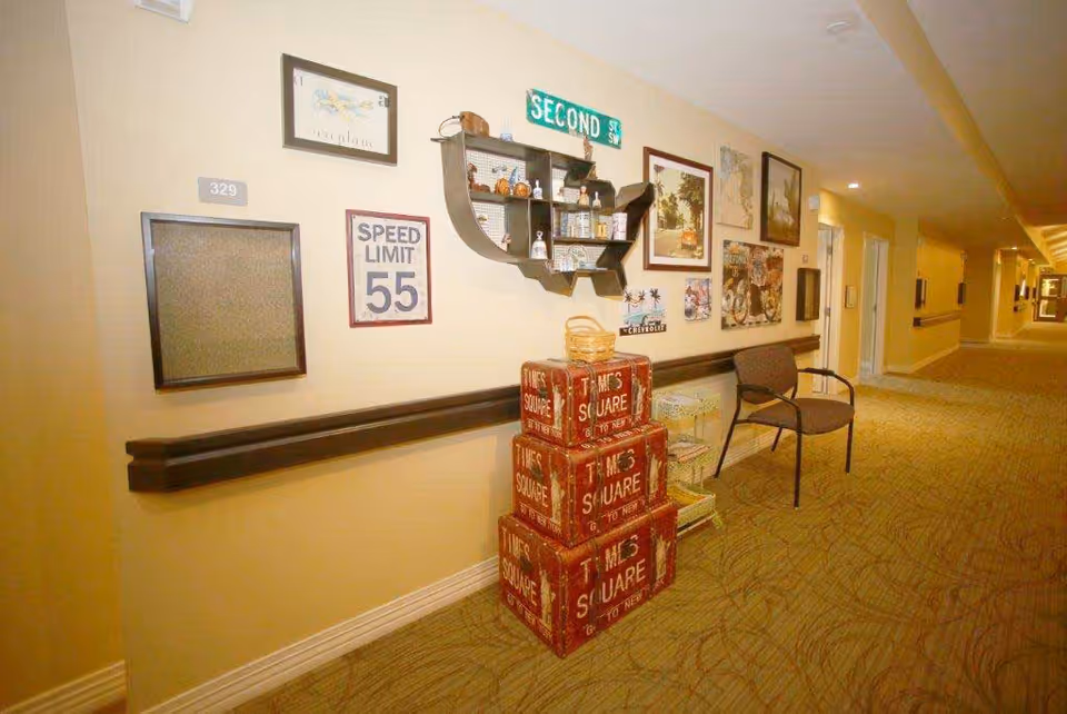 A hallway in a senior living facility with beige walls and carpeted floor. The wall is decorated with various framed pictures, a speed limit 55 sign, a green street sign reading 'SECOND ST SW', and a shelf shaped like the United States holding small decorative items. There are three red vintage-style storage boxes stacked on the floor labeled 'TIMES SQUARE'. A single chair and a small side table are placed along the wall. The hallway extends into the distance with more doors and lighting visible.