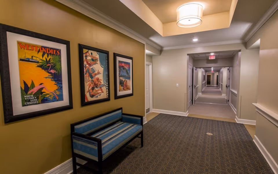 Interior hallway of a senior living facility with beige walls and carpeted floor. There is a blue cushioned bench against the left wall beneath three framed vintage travel posters. The hallway extends into the distance with several doors on either side and a ceiling light fixture above.