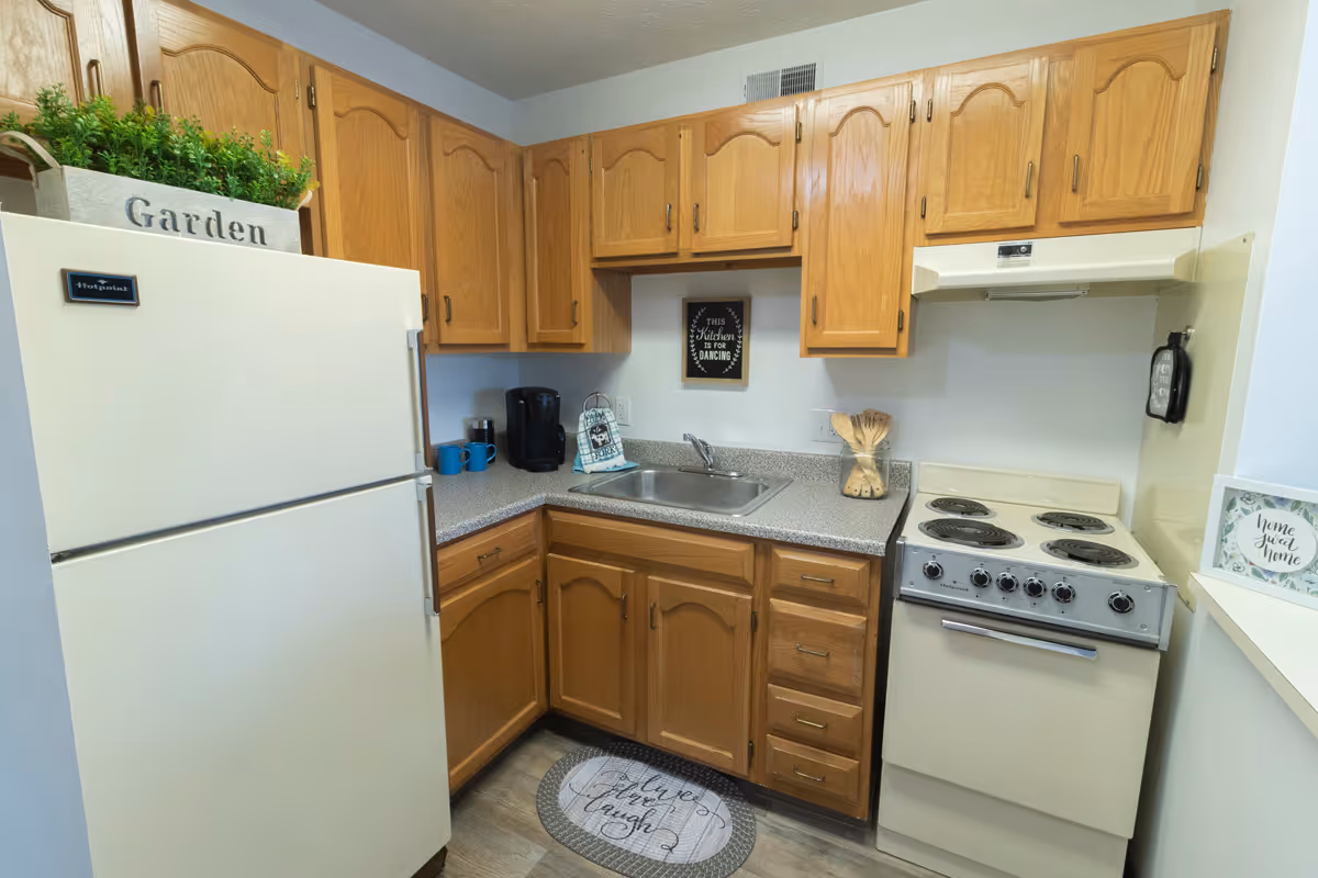 A small kitchen with wooden cabinets, a white refrigerator with a planter labeled 'Garden' on top, a countertop with a coffee maker and mugs, a stainless steel sink, and a white electric stove. There is a small framed sign above the sink that says 'This kitchen is for dancing' and a round floor mat in front of the sink with the words 'Live, Love, Laugh'.