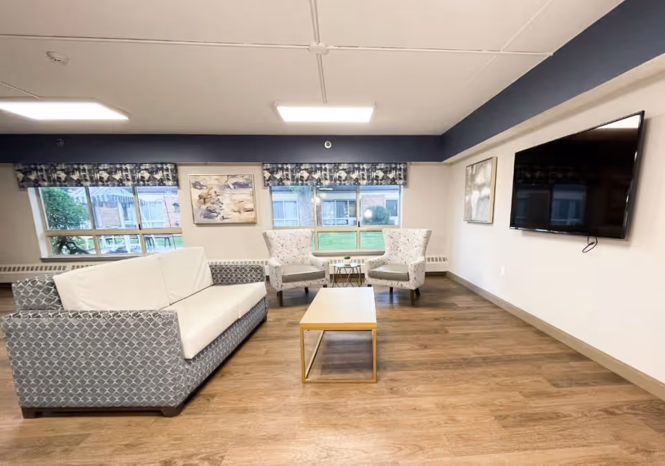 A bright and clean living room area with a patterned gray and white sofa, two matching armchairs, a wooden coffee table, and a large flat-screen TV mounted on the wall. The room has large windows with patterned valances, light-colored walls, and wood flooring.