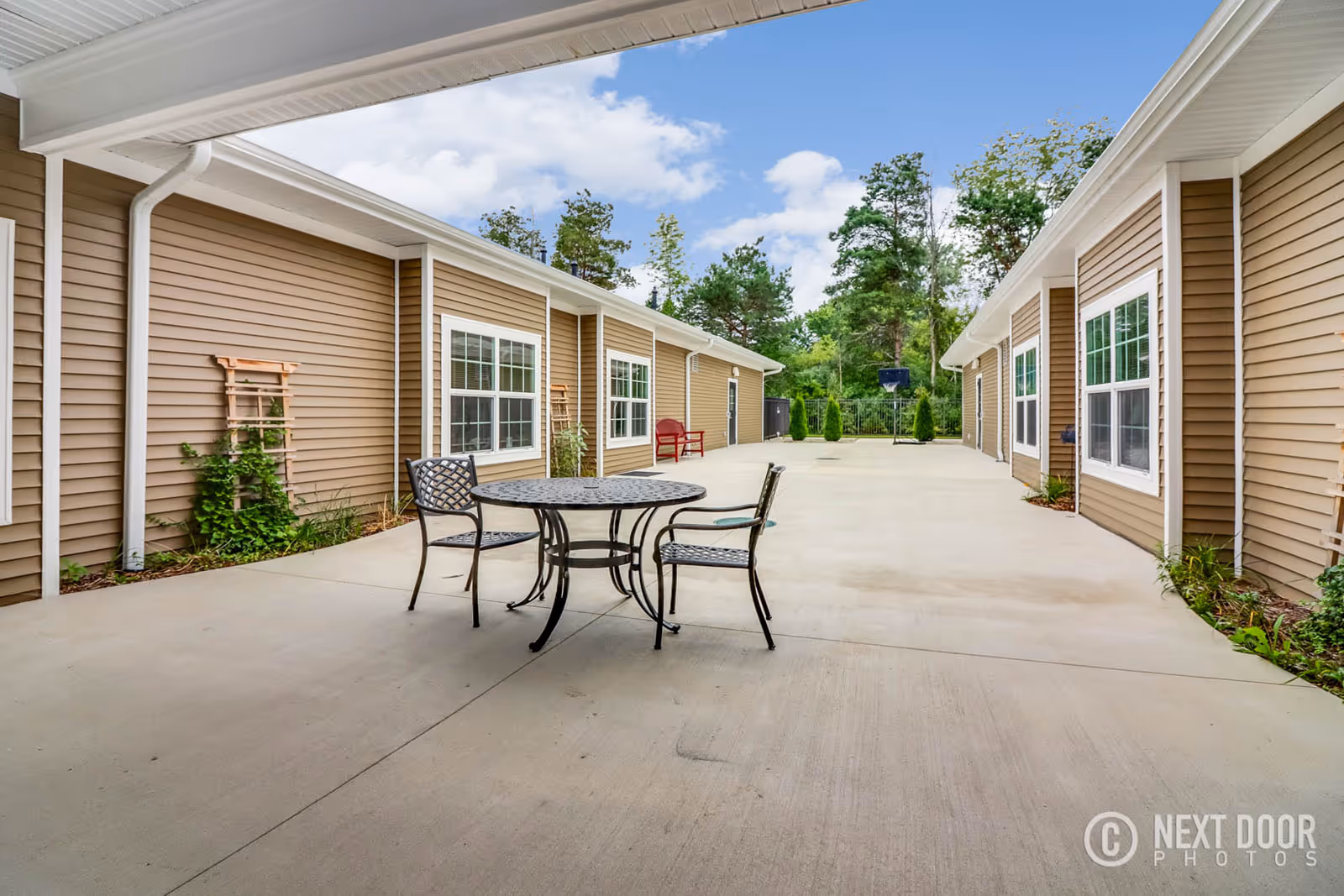 Outdoor courtyard area between two beige buildings with white trim, featuring a round metal table with two chairs on a concrete patio. There is a red bench further back and a basketball hoop near a fence with trees in the background under a partly cloudy sky.
