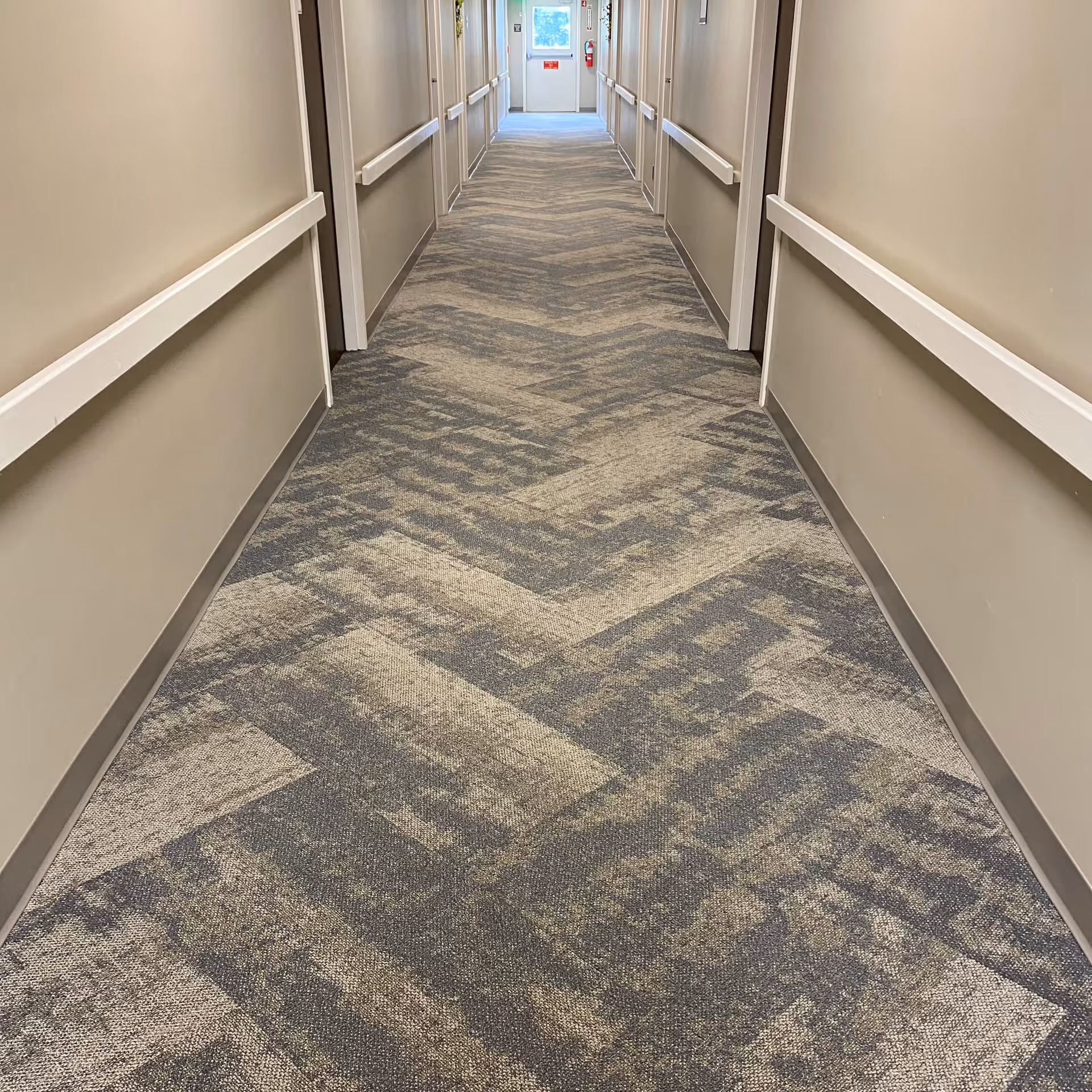A long indoor hallway with patterned carpet flooring and beige walls. The hallway has handrails on both sides and several doorways along the walls. At the end of the hallway, there is a door with a window and a fire extinguisher mounted on the wall.