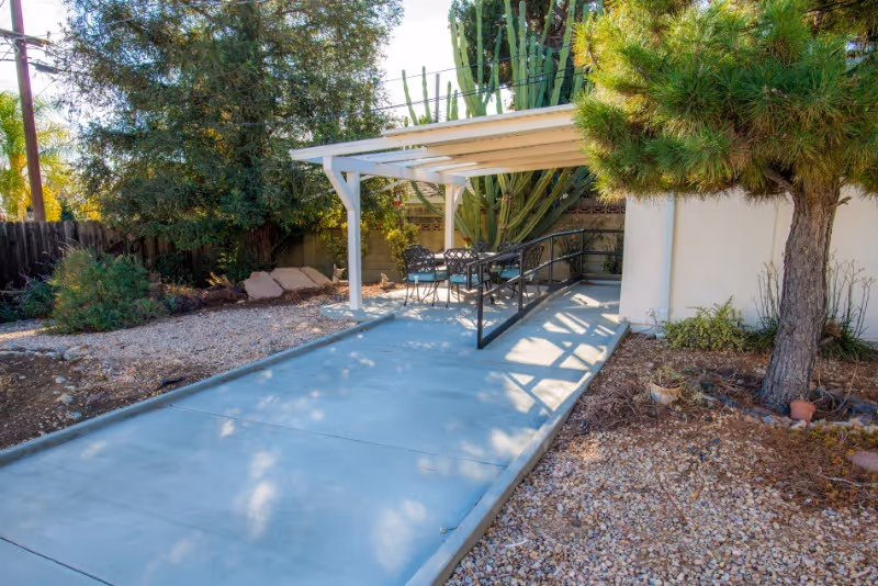 Concrete wheelchair-accessible ramp leading to a covered patio with a table and chairs, surrounded by desert landscaping, a tree, and tall cacti.