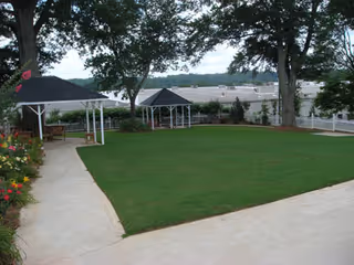 A well-maintained outdoor garden area with a green lawn, two white gazebos, a concrete pathway, colorful flowers on the left side, and large trees providing shade.