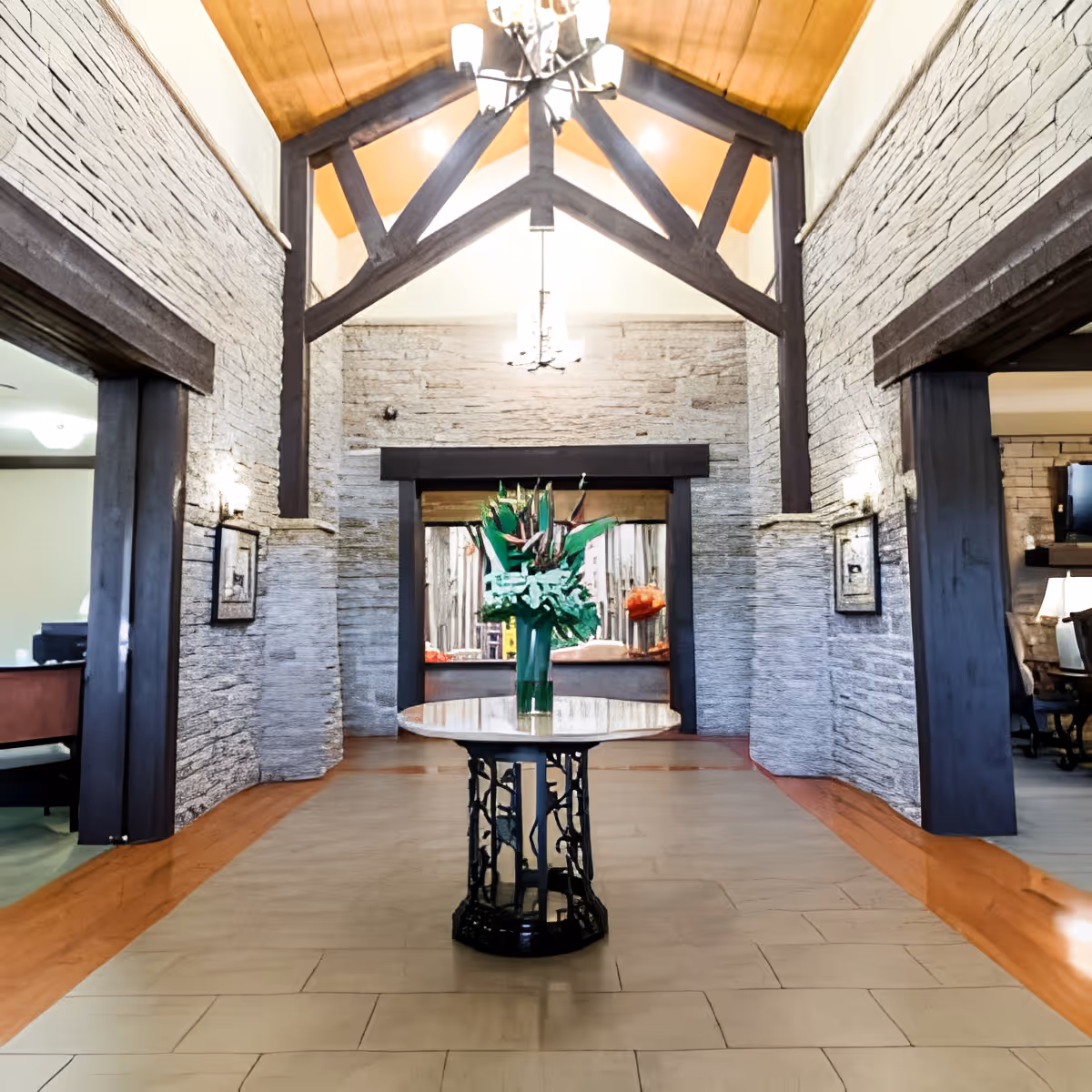 Interior view of a spacious room with high vaulted wooden ceiling and exposed dark wooden beams. The walls are textured with light-colored stone. A round table with a decorative black metal base is centered in the room, holding a tall vase with green and orange floral arrangement. Two doorways on either side lead to other rooms with visible furniture and lighting.