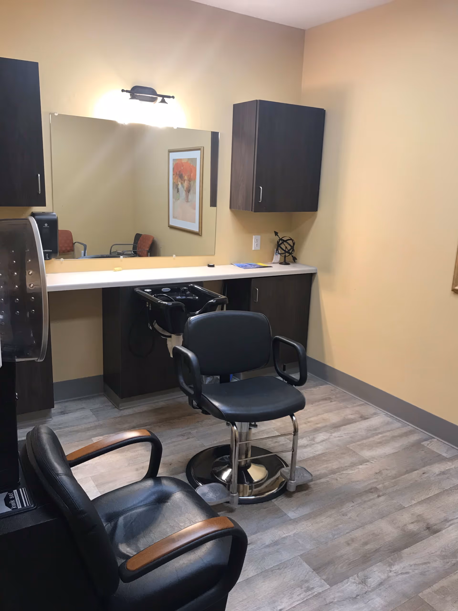 Interior view of a small salon area with two black salon chairs, a large mirror on the wall, a sink for washing hair, dark wood cabinets, and a framed floral picture reflected in the mirror. The walls are painted light yellow and the floor has a wood-like finish.