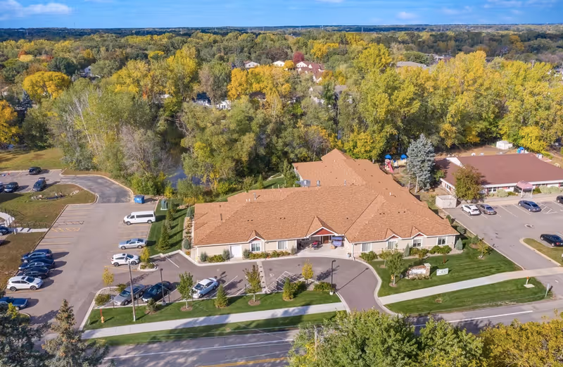 Aerial view of a single-story senior living facility with a central driveway, parking lot, and surrounding trees.