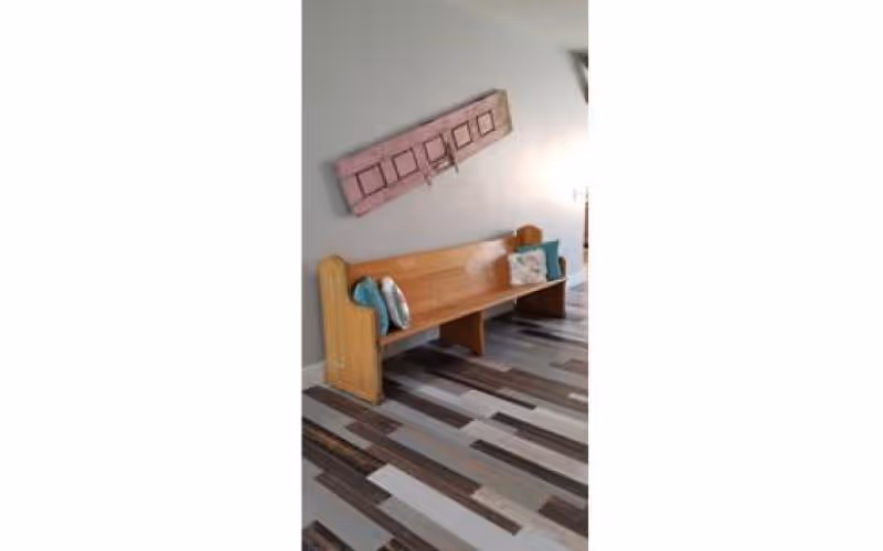 A wooden bench with two decorative pillows placed against a light gray wall. Above the bench, there is a tilted rectangular wall decoration with a pinkish hue and square patterns. The floor has a modern design with alternating light and dark wood planks.