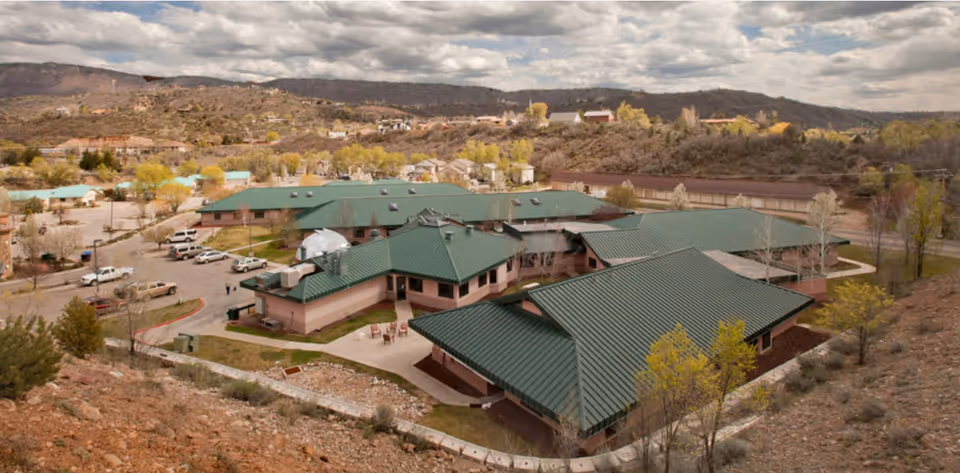 Aerial view of a senior living facility complex with green metal roofs, parking lot, and surrounding hills.